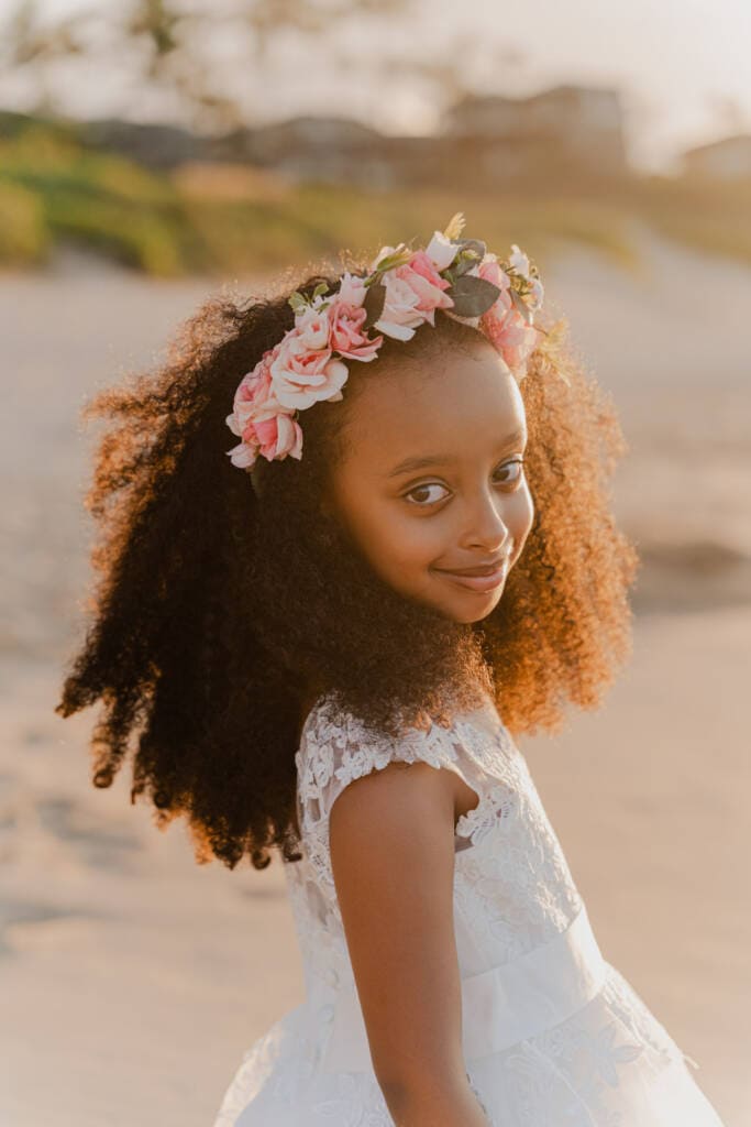 Young girl in a white lace dress wearing a pink floral crown smiling over her shoulder on a Maui beach at golden hour