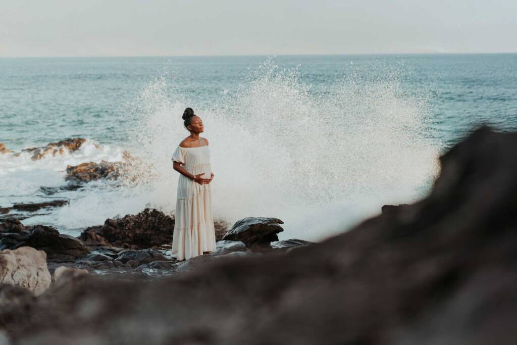 Pregnant woman in a white off-shoulder dress cradling her belly on rocky coastline with a large wave crashing behind her, Maui Hawaii