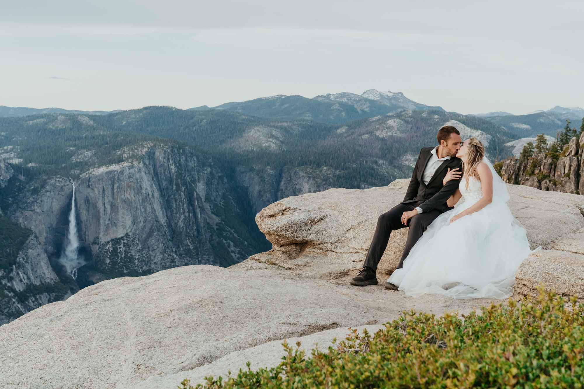 Bride and groom kissing on a granite cliff at Yosemite with Yosemite Falls in the background