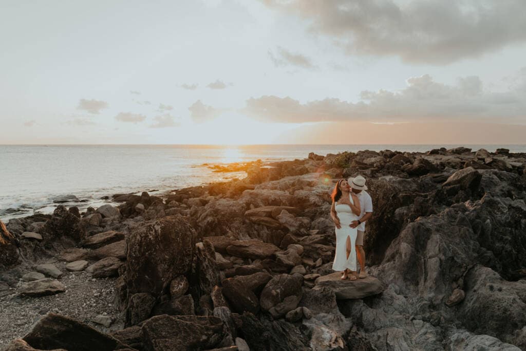 Couple embracing on a dramatic lava rock coastline at sunset, with the ocean and golden sky stretching behind them, Hawaii