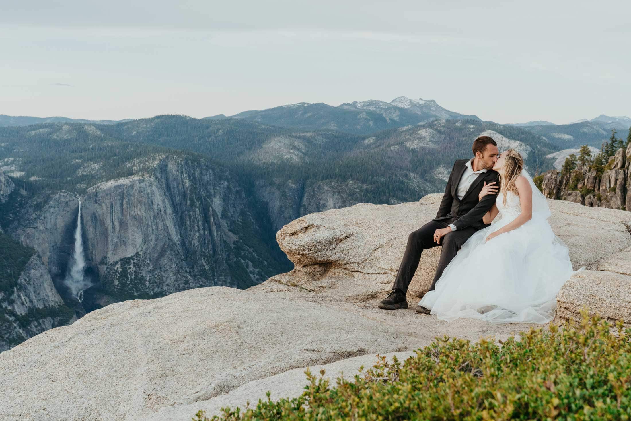 Bride and groom kissing on a granite cliff at Yosemite with Yosemite Falls in the background