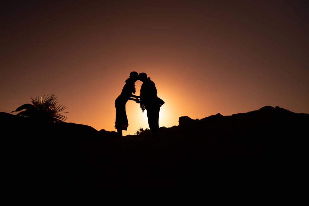 Silhouette of a couple wearing Hawaiian leis kissing at sunset on a rocky hilltop