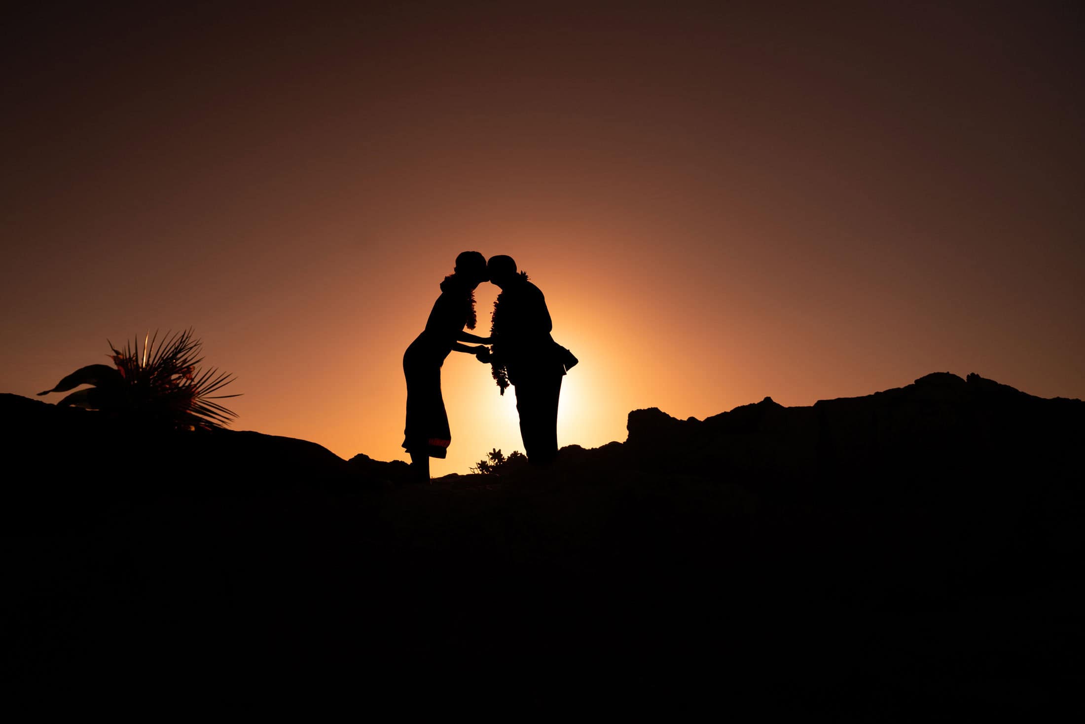 Silhouette of a couple wearing Hawaiian leis kissing at sunset on a rocky hilltop